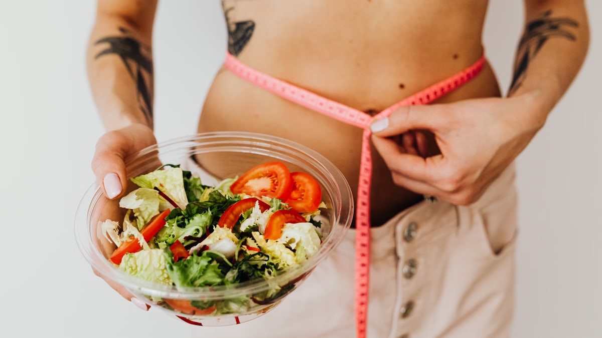 Woman holding vegetable salad and measuring waist with tape, symbolizing healthy lifestyle choices.